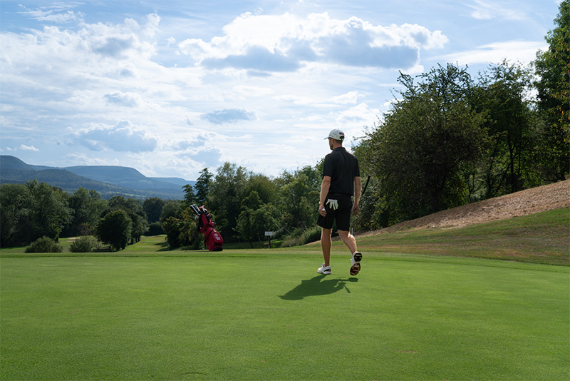 Ein Golfer läuft mit einem Ball in der einen und einem Schläger in der anderen auf eine rote Golfbag zu. Der Golfer ist von hinten zu sehen.