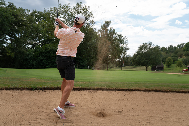 Ein Golfer steht in einer Sandgrube. Man sieht von hinten wie er gerade einen Schlag ausgeführt hat. Der Ball fliegt gerade aus der Grube zusammen mit etwas Sand.