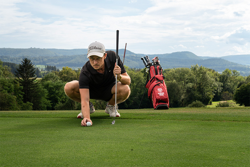 Ein Golfspieler platziert in der Hocke hochkonzentriert einen Golfball auf dem gepflegten grün, um seinen nächsten Schlag erfolgreich durchführen zu können.