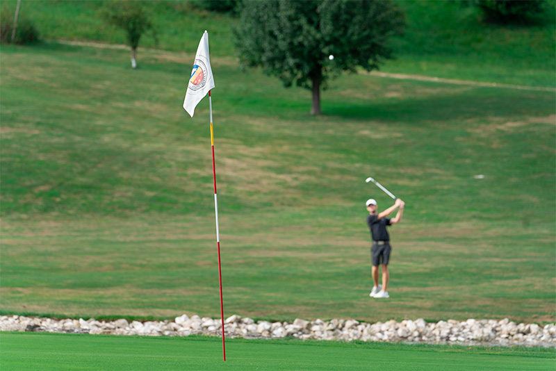 Eine Flagge markiert das Loch auf dem Golfgrün im Vordergrund. Ein Spieler steht mit geschwungenem Schläger im Hintergrund und beobachtet seinen Schlag.