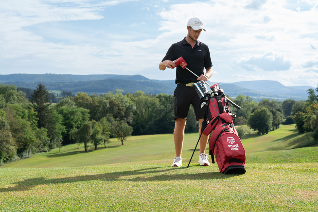 Ein Golfer steht auf dem Grün und packt seinen Schläger in eine rote Golfbag ein, die neben ihm steht.